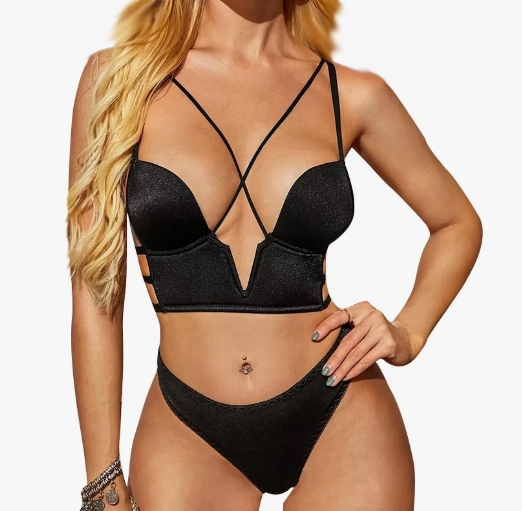 Woman in a black strappy lingerie set with crisscross straps, high-cut bottoms, blonde hair, and a belly button piercing with hand on hip.